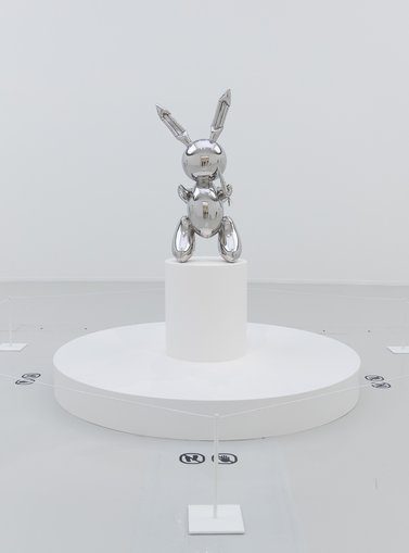 A stainless steel sculpture of a rabbit in the style of a balloon figure, placed atop a white pedestal in a gallery