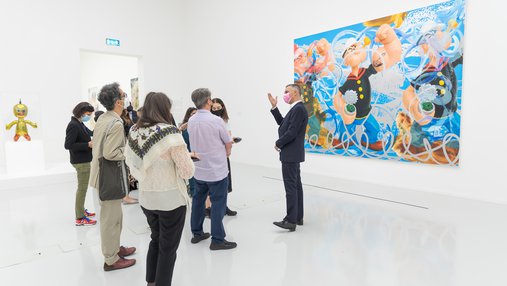 Massimiliano Gioni talking to a group of visitors in a gallery, standing in front of Jeff Koons' painting Triple Popeye