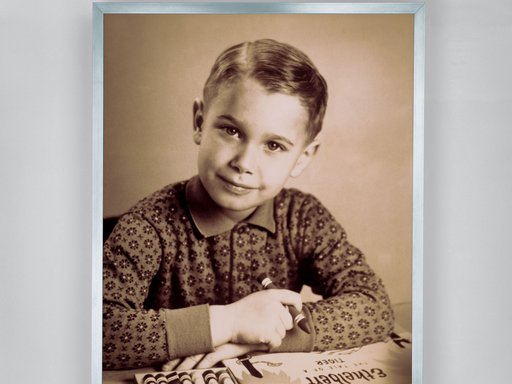A black-and-white photograph of Jeff Koons as a child sitting at a table with a box of crayons and holding a large crayon in his right hand