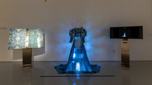 A dress installation that lights up in blue, a TV showing a video about the dress is on the right and two paintings displayed on the left.