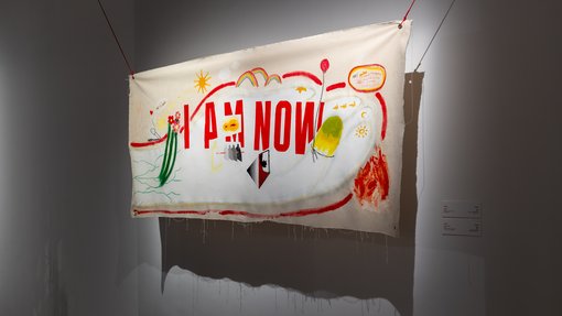 A piece of fabric suspended in the air with the sentence 'I Am Now' written on it.