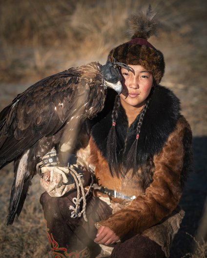 A child from the central desert wearing a traditional dress and holding a large blindfolded falcon in her hand
