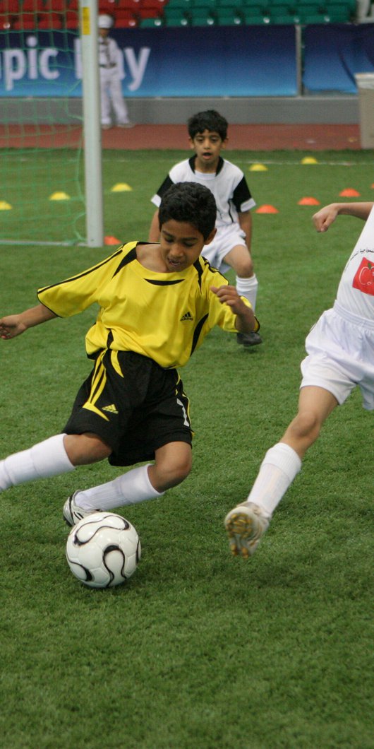 Two young boys in yellow jerseys and three in white jerseys go after a football while a goalie in an orange uniform looks on.