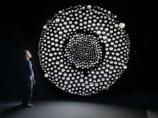 A man standing in a dark room facing a large circle of dozens of round, backlit mirrored shapes