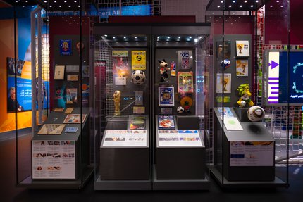 Four glass exhibition cases, each displaying text/photo content, footballs, soft toy mascots and a model of the Jules Rimet FIFA trophy.