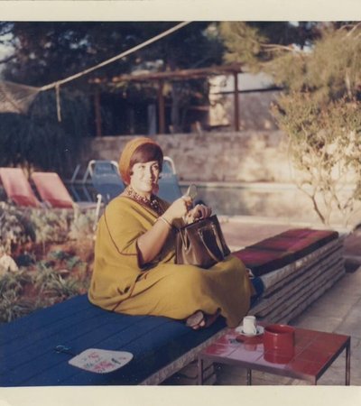 A photograph of a woman wearing a caftan sitting on a bench outside, holding her bag on her lap.