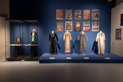 A gallery showcasing Palestinian dresses embroidered with a symbol of the Palestinian flag with posters in the back depicting war times in Palestine
