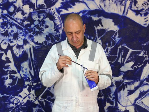 Visiting Artist Nedim Kufi standing in front of his painting, in his studio at the Fire Station, with oil paint and paint brush in hand.