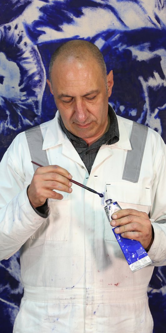 Visiting Artist Nedim Kufi standing in front of his painting, in his studio at the Fire Station, with oil paint and paint brush in hand.