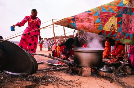 امرأة ترتدي ثوباً ملوناً أمام خيمة ملونة ويظهر في الصورة قدر طعام على النار