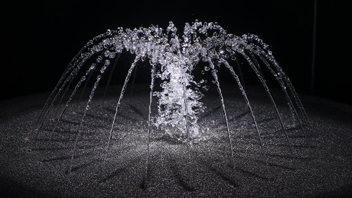 A small water fountain in a dark room, the fountain is lit up by white lights.