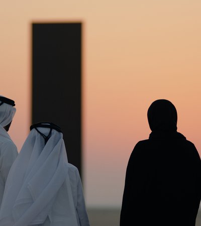 Three people in a desert landscape looking off into the sunset and two large black monoliths in the background.