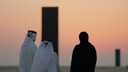 Three people in a desert landscape looking off into the sunset and two large black monoliths in the background.