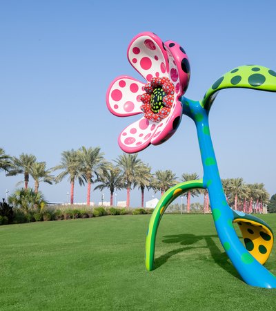 Sculpture of a big flower in multiple colours sits on a grassy hill, palm trees in the background.