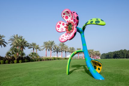 Sculpture of a big flower in multiple colours sits on a grassy hill, palm trees in the background.