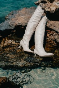 A pair of high-heeled white leather women's boots positioned on rocks by the sea.