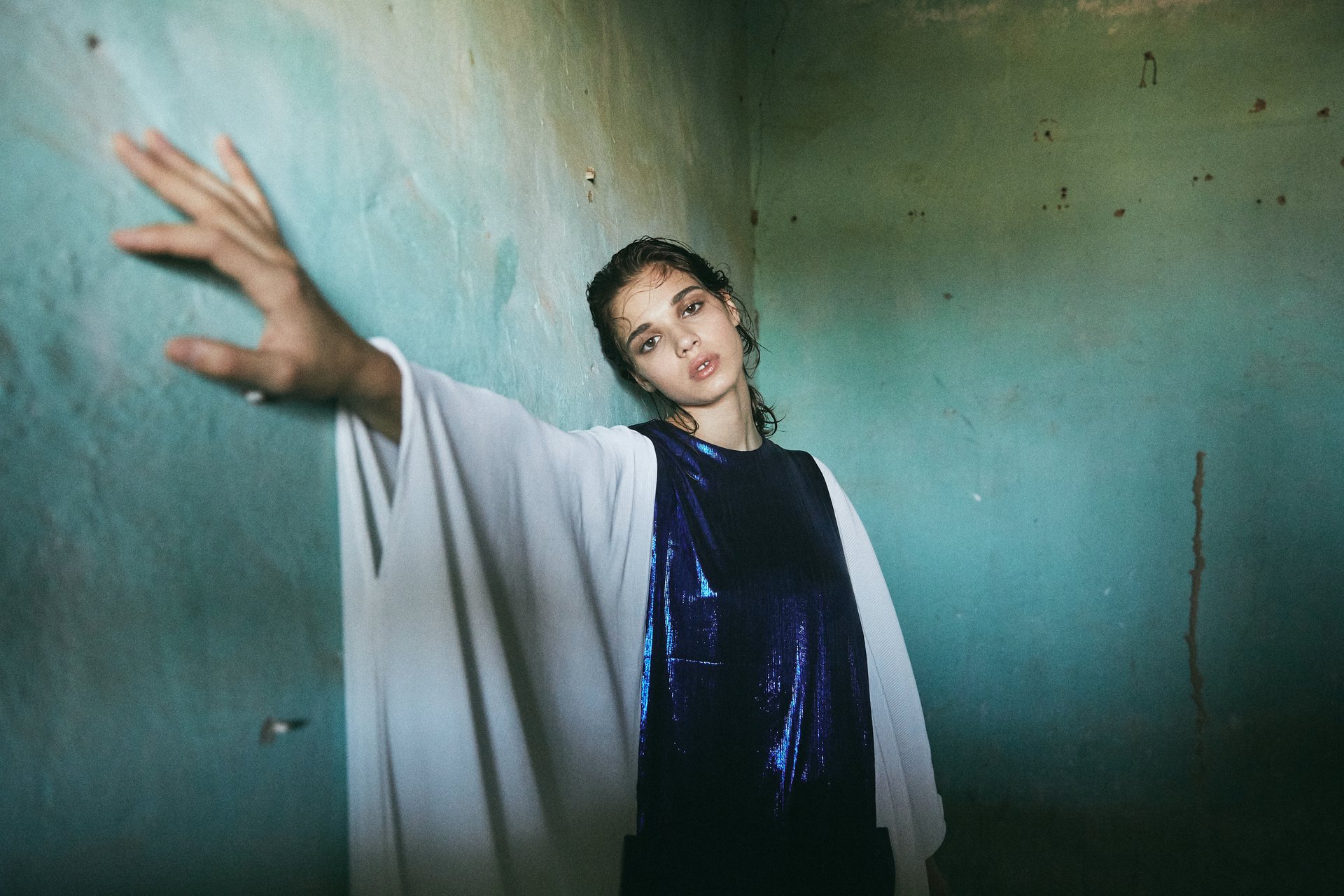 Woman standing against a wall with her arm outstretched, wearing a blue dress and abaya.