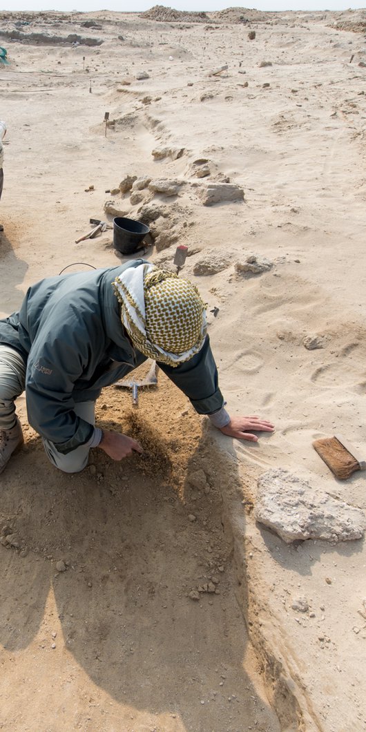 Two men kneeling on sandy desert ground using small tools to expose archeological remains at Al Zurbarah, Qatar