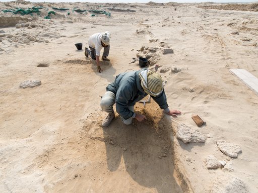 Two men kneeling on sandy desert ground using small tools to expose archeological remains at Al Zurbarah, Qatar