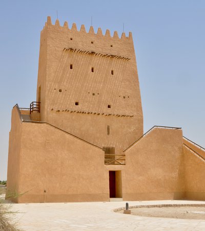 A tall straight-sided sand coloured building with a doorway, external staircase to upper doors, small windows and a spiked roofline