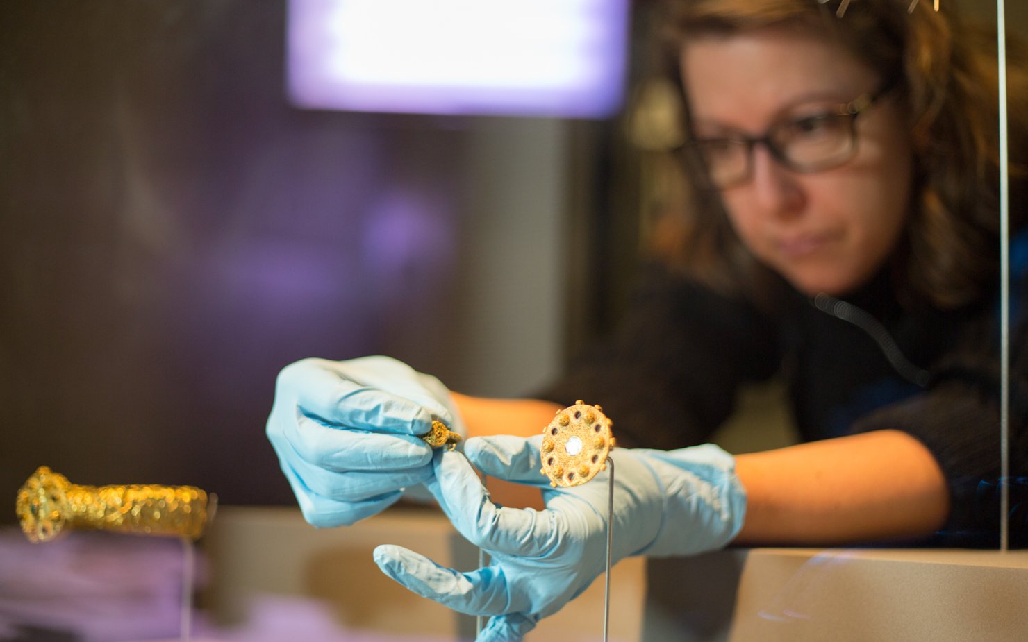 A woman wearing gloves concentrates on placing a small gold jewel inside a glass display