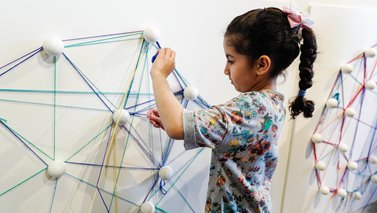 A young child making geometric shapes on a wall with string, at Dadu, Children's Museum