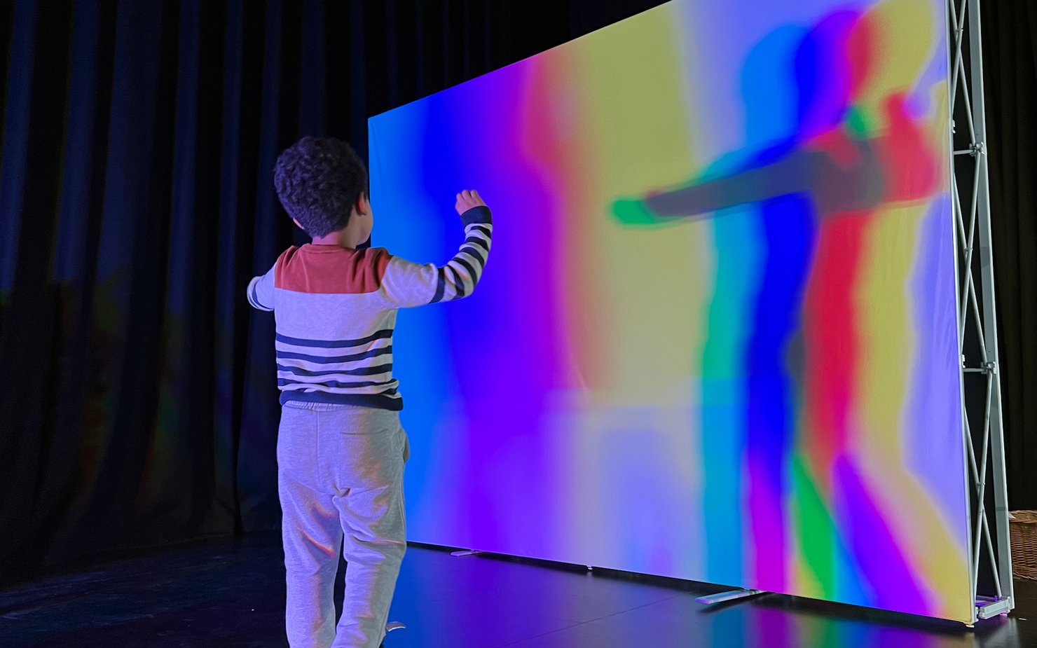 A boy standing in front of a white screen reflecting his shadow in various colour