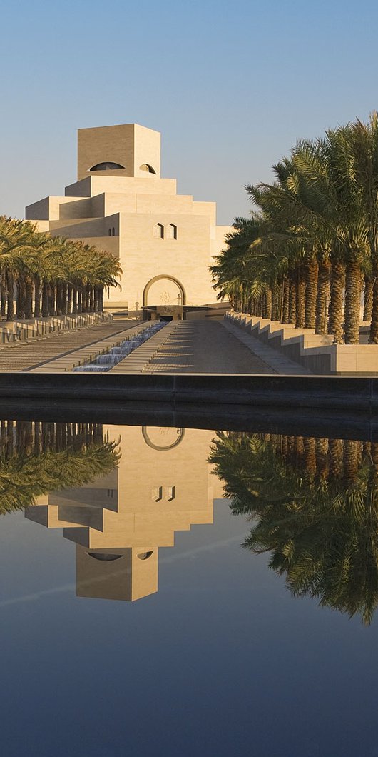 مجموعة من أشجار النخيل محيطة بجانبي المدخل الرئيسي لمتحف الفن الإسلامي وتبدو واجهة المتحف منعكسة على سطح الماء