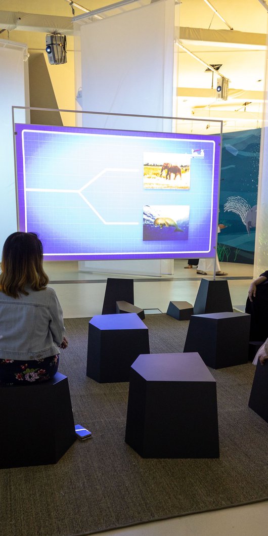 Parents and child sitting on stools with a tour guide watching a video on screen