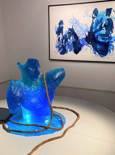 A blue sculpture of a man in front of a blue painting depicting a tribe