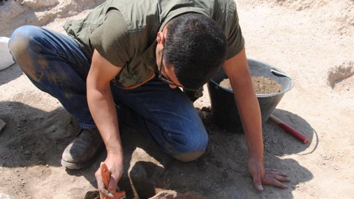 Hatem Arrok bending down and holding a brush while excavating fossils in a desert