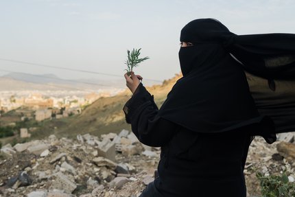 A woman dressed in black and wearing a veil holds a green sprig in her hand.
