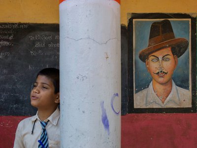 A young boy leans against a concrete column with a striped wall and painting of a man with a hat in the background.