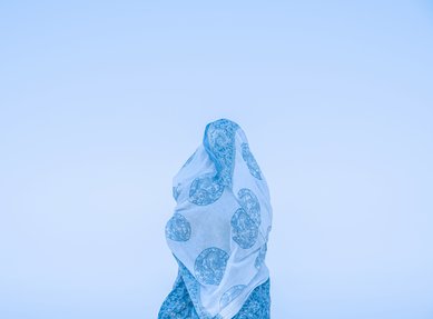 A photo of a female with white and blue fabric wrapped around them standing against a light blue background.