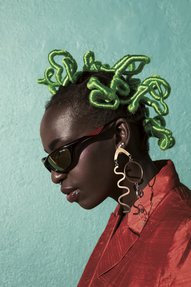 A black woman stand in profile, wearing her hair wrapped in bright green threat, sunglasses, long earrings and a red silk jacket.