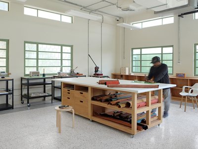 Interior view of a workroom with a central bench and a man crafting with leather.
