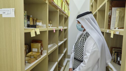 A visitor in traditional Qatari Thobe looking at archival manuscripts and books stacked in library shelves