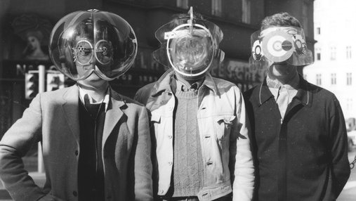 Black and white picture of three men wearing futuristic face shields and covers. Scene is from the mid- 20th century, men are wearing jackets.