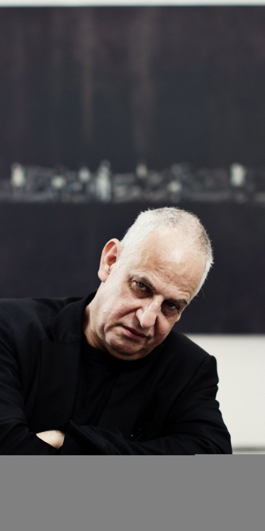 A portrait of Luc Tuymans framed by a black artwork in a museum while looking directly at the camera.