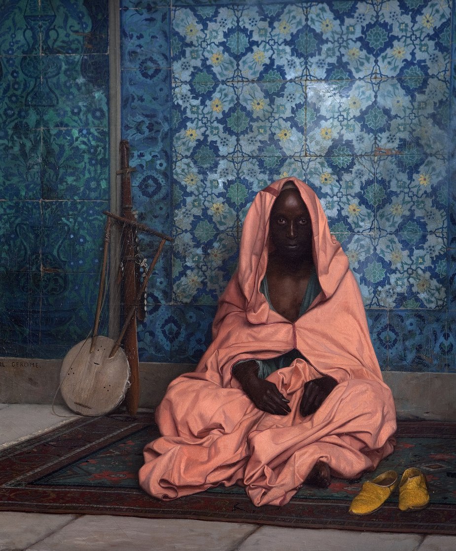 A man wrapped in pink fabric sits cross legged on a carpet, a musical instrument in the background.