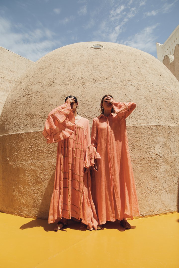 Two women standing in front of a small domed building, wearing long pink dresses.