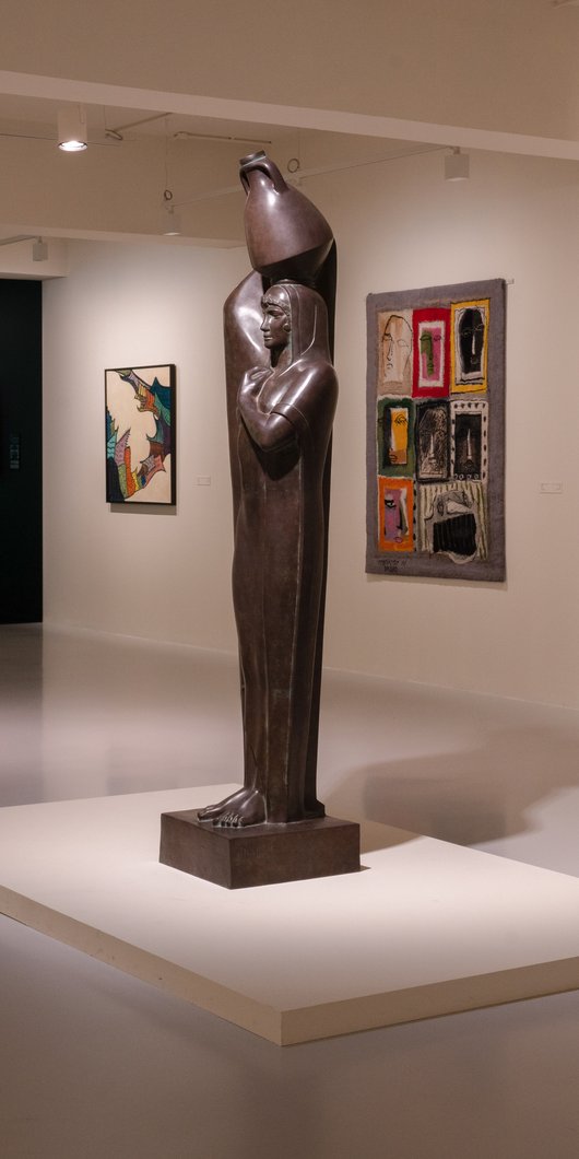 Photo of a gallery space with a sculpture of a peasant women with a jug on her head in the middle.