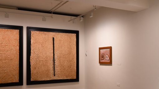 A corner of an exhibition gallery, one side with large black/sand framed paintings and one with a small brown framed pink hued work.