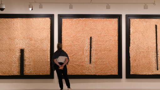 Woman viewing three large, framed, square artworks, textured to look like straw, with black lines.