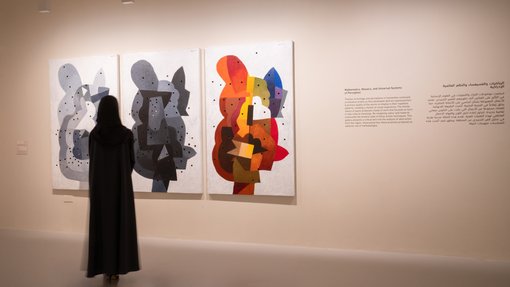 A woman in an abaya viewing three abstract artworks in a gallery space; one grey, the next darker and the third in colour.