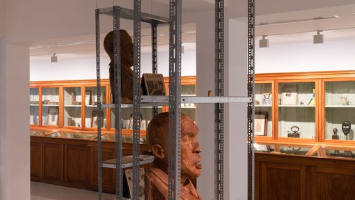 Gallery view of a large glass and wooden case with items displayed within and two rust-coloured busts in the foreground sitting on a metal case.