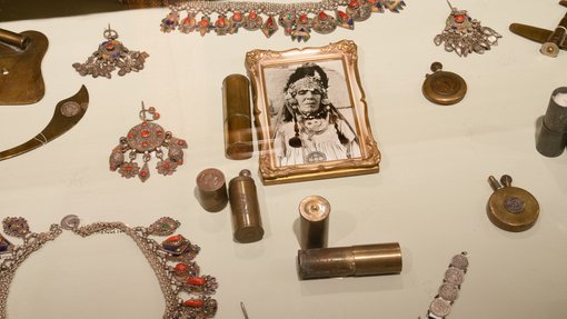 Brass and gold-hued jewellery displayed on a white background surrounding a black and white framed photo of a woman in traditional dress.