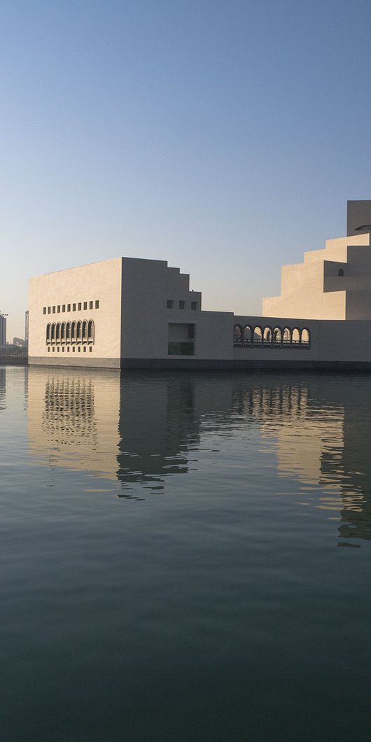An exterior view of the Museum of Islamic Art during the day, showing the building's reflection in the water