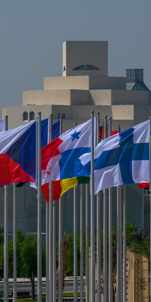 International flags on high poles with a geometric, pyramid-like building in the background.