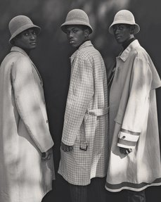 Three stylish young black men stand together wearing tailored rain coats and hats.